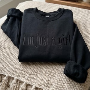 Może przedstawiać: Czarna bluza z haftowanym napisem "I'm just a girl". Bluza z okrągłym dekoltem ma długie rękawy z podwiniętymi mankietami. Bluza jest prezentowana na beżowej, teksturowanej powierzchni.