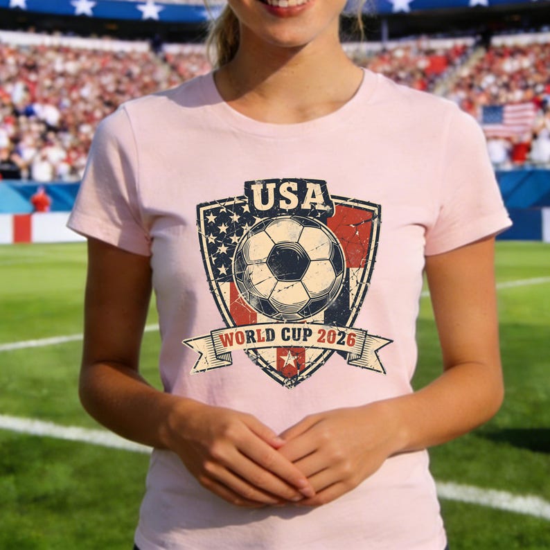 Puede incluir: Camiseta rosa claro con un gr&aacute;fico desgastado. El gr&aacute;fico es un bal&oacute;n de f&uacute;tbol dentro de un escudo con el texto "USA WORLD CUP 2026". El dise&ntilde;o del escudo incorpora las estrellas y rayas de la bandera estadounidense.