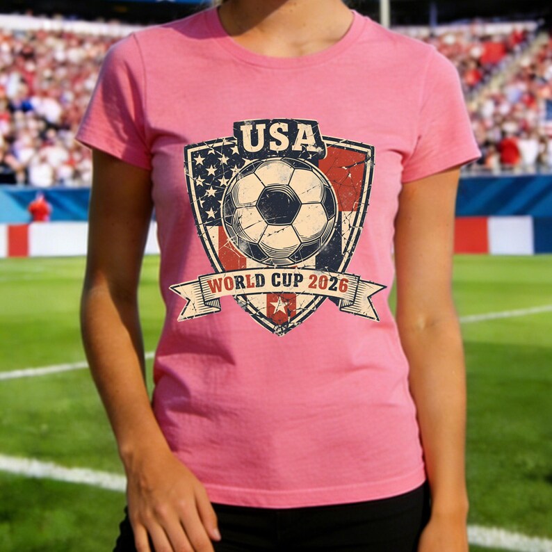 Puede incluir: Camiseta rosa con un gr&aacute;fico desgastado de un bal&oacute;n de f&uacute;tbol dentro de un escudo. El escudo incorpora la bandera estadounidense y el texto "USA" y "WORLD CUP 2026". La camiseta es de cuello redondo.