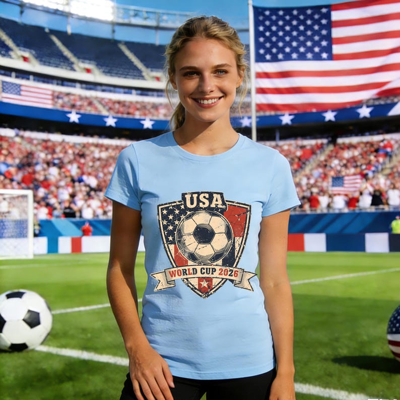 Puede incluir: Camiseta azul claro con un dise&ntilde;o gr&aacute;fico que incluye un bal&oacute;n de f&uacute;tbol, la palabra "USA" y "World Cup 2026" en un escudo. La imagen est&aacute; tomada en un estadio con una bandera americana de fondo.