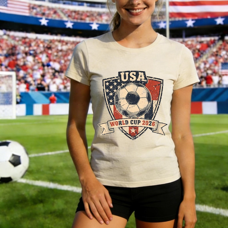 Puede incluir: Camiseta beige con un dise&ntilde;o de bal&oacute;n de f&uacute;tbol dentro de un escudo, con el texto "USA" y "World Cup 2026". El dise&ntilde;o incorpora las estrellas y rayas de la bandera estadounidense. La camiseta se lleva en un campo de f&uacute;tbol.