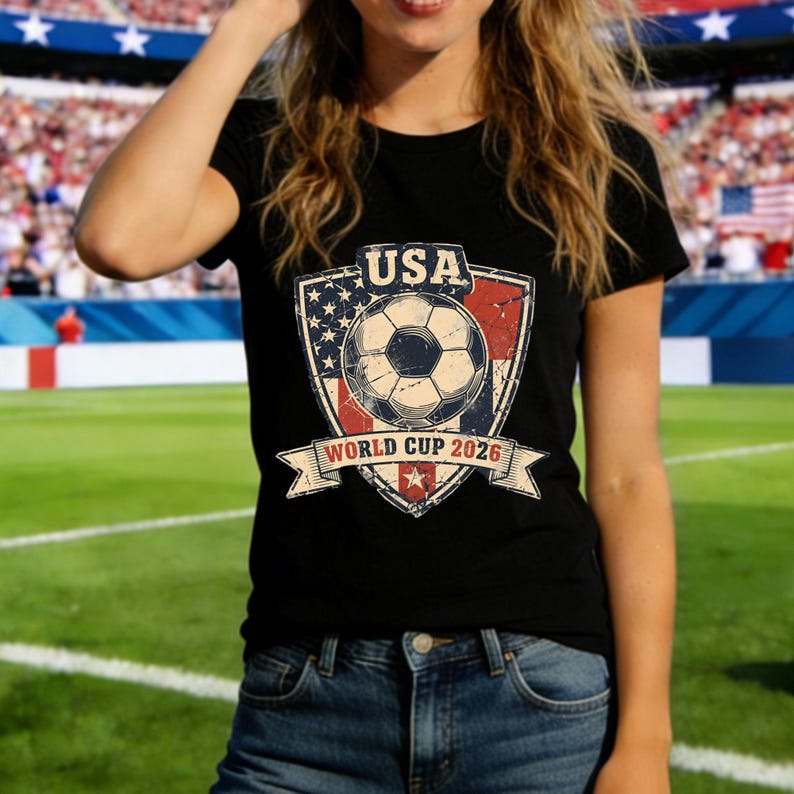 Puede incluir: Camiseta negra con un dise&ntilde;o de bal&oacute;n de f&uacute;tbol, el texto "USA" y "World Cup 2026". El dise&ntilde;o utiliza los colores de la bandera estadounidense y tiene un aspecto vintage. La camiseta la lleva una persona en un estadio.