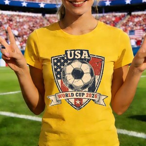 Puede incluir: Camiseta amarilla con un gr&aacute;fico desgastado de un bal&oacute;n de f&uacute;tbol dentro de un escudo con el texto "USA" y "World Cup 2026". El dise&ntilde;o del escudo incorpora la bandera estadounidense. La camiseta es de manga corta.