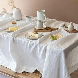 May include: A white linen tablecloth covers a wooden table, set with various white ceramic dishware, including a mug, a creamer, and small plates holding food items like cheese, grapes, and butter. Dried plants and small vases add to the aesthetic.