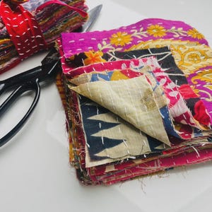 Peut inclure: Un ensemble de carrés de tissu colorés avec des motifs floraux et géométriques, et un paquet de tissu similaire attaché avec un ruban rouge. Des ciseaux noirs sont placés à proximité. Les tissus sont dans des tons de rose, jaune, bleu et rouge.