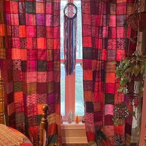 May include: Patchwork curtains in shades of red, pink, and orange, with a variety of patterns. A dreamcatcher hangs in the window. The curtains are made of fabric squares sewn together, creating a bohemian style.