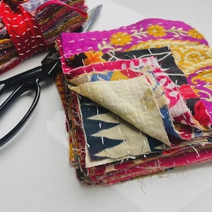 May include: A stack of colorful, patchwork fabric squares with floral and geometric patterns. The squares are in shades of pink, yellow, blue, and red. A pair of black scissors and a bundle of fabric squares tied with a red ribbon are also present.