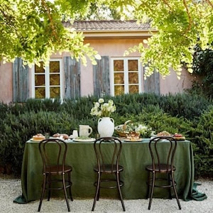 May include: An outdoor dining setting featuring a green tablecloth, three dark wooden chairs, and a table laden with food and flowers. The scene is set in a garden with a pink building and lush greenery in the background.
