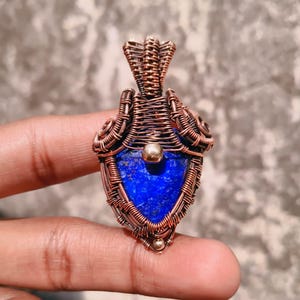 Può includere: Un ciondolo fatto a mano con una vibrante pietra blu incastonata in una cornice di filo di rame. L'intricato lavoro a filo circonda la pietra, creando un design unico. Il ciondolo è tenuto dalle dita di una persona.