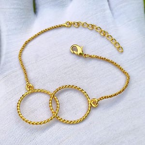 Op de afbeelding: Een goudkleurige armband met twee in elkaar grijpende cirkelvormige ringen van gedraaid metaaldraad. De armband bevat een fijn kettingsegment met een karabijnsluiting en een verstelbare verlengketting met kleine schakels.