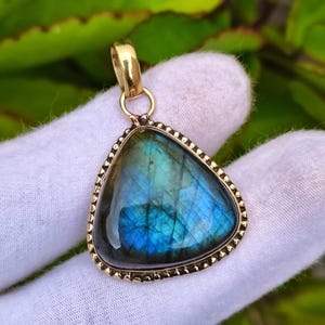 May include: A triangular labradorite pendant featuring a polished, iridescent blue and green gemstone set in a gold-toned metal frame with a beaded edge and a simple gold-toned bail at the top for attachment to a necklace chain or cord.