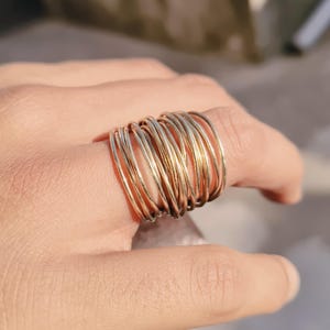 May include: A gold ring featuring numerous thin, intertwined bands. The ring is displayed on a finger, highlighting its detailed design and metallic lustre. The bands create a layered effect, reflecting light and adding visual interest to the jewellery.