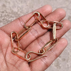 May include: A gold-coloured chain bracelet with a distinctive design. The bracelet comprises hammered links and safety pin-shaped clasps. The bracelet is held in a hand, highlighting its intricate details and warm metallic hue.