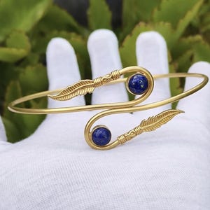 Peut inclure: Un bracelet de manchette doré avec un motif de plume à chaque extrémité. Le bracelet comporte deux pierres précieuses rondes bleu foncé. Le bracelet est présenté sur une surface blanche avec un fond de feuillage vert.