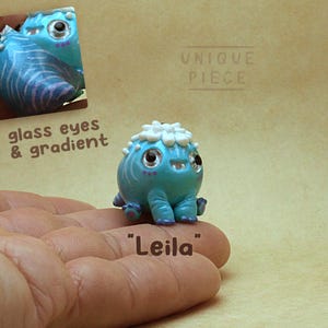 Könnte beinhalten: Eine kleine, runde, türkisfarbene Kreaturenfigur namens Leila mit einem weißen blumenartigen Kamm auf dem Kopf, glänzenden Glasaugen und violetten Akzenten. Der Text UNIQUE PIECE, glass eyes & gradient und Leila ist im Bild zu sehen.