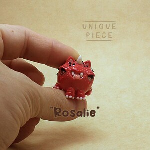 Peut inclure: Une petite figurine de créature rouge avec une corne blanche unique, plusieurs yeux noirs et des dents visibles. La figurine a une surface texturée et porte l'inscription "UNIQUE PIECE" ainsi que le nom "Rosalie" en lettres brunes.