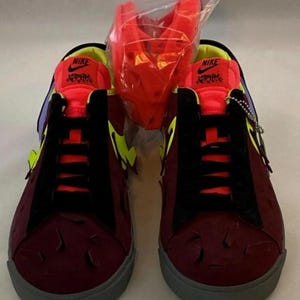 May include: A pair of burgundy sneakers with black accents, yellow and purple details, and gray soles. The sneakers have the Nike logo on the tongue and a textured design on the toe. A red accessory is in a clear plastic bag.