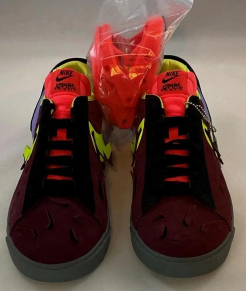 May include: A pair of burgundy sneakers with black accents, yellow and purple details, and gray soles. The sneakers have the Nike logo on the tongue and a textured design on the toe. A red accessory is in a clear plastic bag.