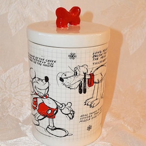 May include: A white ceramic jar with a lid and a red bow-shaped handle. The jar features illustrations of Mickey Mouse and Pluto, with text and grid lines. A decorative item for the kitchen.