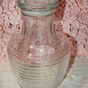 May include: Clear glass jar with a lid. The jar has a rounded base with horizontal ribbed detailing and a narrower neck. The lid is also glass and fits snugly on top. The jar is likely for storage or display.