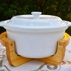 May include: A white ceramic casserole dish with a matching lid and integrated side handles, resting securely within a light-toned wooden cradle stand. The smooth, glossy finish of the pot contrasts with the natural grain of the wooden base.