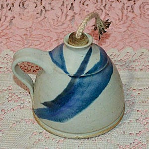 May include: A handmade ceramic oil lamp with a white and blue design. The lamp has a handle and a lid with a wick. The lamp is sitting on a pink lace tablecloth.