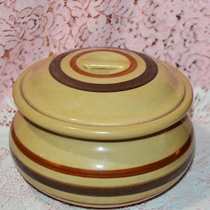 May include: A ceramic lidded jar with a light yellow base and lid. The lid features concentric brown and orange bands, with a small handle. The jar's body has similar bands. The jar is likely for kitchen or home decor.