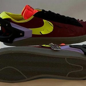 May include: Burgundy and black sneakers with a yellow Nike swoosh. The shoes have a unique design with a black heel counter, a purple accent, and a gray sole. The tongue is orange and yellow.