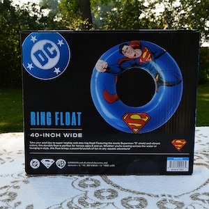 May include: A black product box for a 40-inch wide Superman ring float. The box features a blue DC logo, an illustration of Superman, and the red and yellow Superman shield emblem. Text includes "RING FLOAT" and "40-INCH WIDE" with branding.