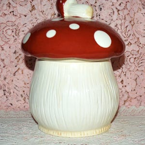 May include: A ceramic mushroom-shaped cookie jar. The lid is a vibrant red with white polka dots, and a small mushroom handle. The base is white with vertical textured lines. The jar is a whimsical kitchen decor item.