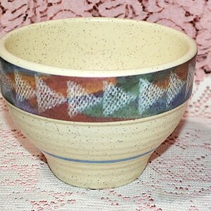 May include: A speckled ceramic bowl with a cream-colored exterior and a decorative band featuring geometric patterns in shades of brown, green, and blue. A blue stripe encircles the base. This handcrafted bowl is suitable for serving.