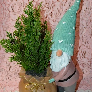 May include: A whimsical garden gnome figurine holding a small potted evergreen plant. The gnome wears a mint green hat with white heart accents, a pink coat, and gray pants. The plant is in a brown bag with a gold bow.
