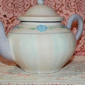 May include: A ceramic teapot with a lid, spout, and handle. The teapot is primarily white with vertical blue stripes and a blue floral design. The spout and handle are a light lavender color. The teapot is sitting on a white lace tablecloth.