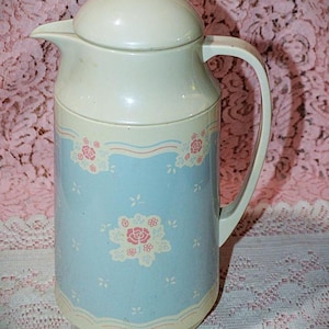 May include: Vintage insulated thermos in a light blue and cream color scheme. The thermos features a floral design with pink and white roses. It has a handle and a lid for pouring.