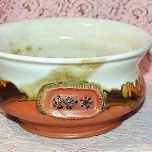 May include: A ceramic bowl with a wide rim and a rounded base. The top is white, transitioning to brown and orange with a decorative rectangular panel featuring floral designs. The bowl is likely handmade.
