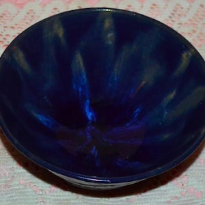 May include: A deep blue ceramic bowl with a glossy finish. The bowl's interior features a swirling pattern of lighter blue and gold hues, creating a dynamic visual effect. The bowl is round and appears to be handmade.