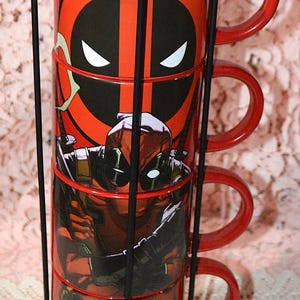 May include: A set of four red ceramic mugs featuring Deadpool graphics, stacked in a black metal rack. Each mug has a handle and a different Deadpool image, with the iconic mask and character poses.