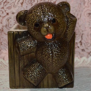 May include: A brown ceramic teddy bear planter with an orange tongue and black eyes. The bear is emerging from a brown wooden crate-style planter. The planter has a textured surface and is suitable for small plants or decorative items.