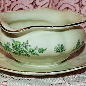 May include: Cream-colored gravy boat and saucer set with a scalloped rim and gold trim. The boat and saucer are decorated with green floral patterns. A classic addition to any dining table.