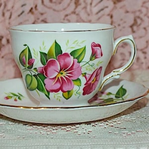 May include: A white teacup and saucer set with a floral design. The cup and saucer are decorated with pink flowers, green leaves, and gold trim. The teacup has a curved handle.