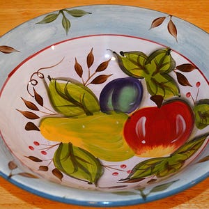 May include: A hand-painted ceramic bowl with a light blue rim and a white interior. The bowl features a colorful fruit design, including a red apple, a yellow pear, and a purple plum, surrounded by green leaves and brown accents.
