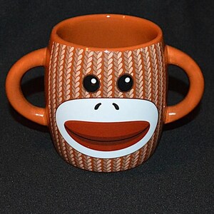 May include: A ceramic mug featuring a brown and tan knit-patterned exterior with two side handles. The front displays a stylized monkey face with large black eyes, a white oval muzzle, and a wide, curved red mouth centered on the mug body.