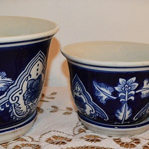 May include: Three ceramic flower pots in varying sizes. The pots are white on the inside and feature a dark blue exterior with white floral and leaf patterns. The pots are arranged on a white lace tablecloth.