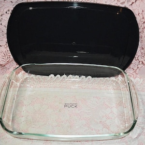 May include: A clear glass rectangular baking dish with rounded handles and the text "WOLFGANG PUCK" in the center. A black lid sits on top. The dish is on a pink floral patterned surface.