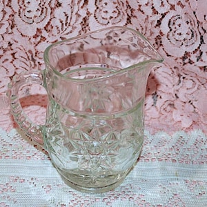 May include: A clear glass pitcher with a handle and a spout. The pitcher has a decorative starburst pattern on the bottom and a ribbed design around the middle. The handle is also textured. The pitcher is likely used for serving beverages.