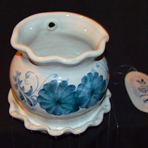May include: White ceramic wall pocket with a scalloped rim and a matching saucer base. The pocket is decorated with blue floral designs. A small tag with a matching floral design hangs from the pocket.