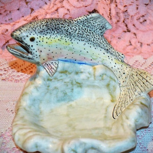 May include: A ceramic dish featuring a sculpted trout fish with a speckled grey and white body, open mouth, and fins. The base of the dish is shaped like a textured, light-colored rock or water formation with mottled green and tan accents.
