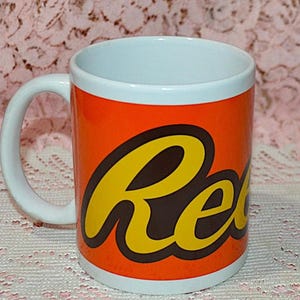 May include: A white ceramic mug with an orange background featuring the Reese's logo in yellow and brown. The mug has a white handle and a white interior. The mug is cylindrical and suitable for hot or cold beverages.