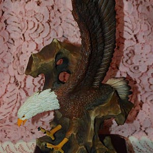 May include: A detailed sculpture of a bald eagle in flight, perched on a weathered tree trunk. The eagle has brown and white feathers, yellow talons, and an orange beak. The base is a dark brown rectangle with a silver plaque that reads "Bald Eagle".