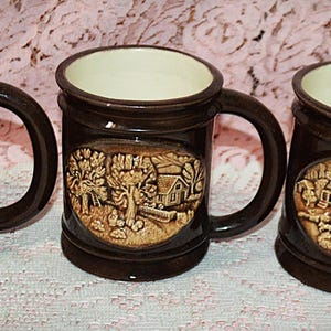 May include: Three brown ceramic mugs with cream-colored interiors and large handles. Each mug features a raised relief scene of a rural landscape with a house, trees, and animals. The mugs are displayed on a pink patterned surface.